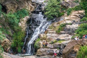 Ein kleiner Wasserfall fließt eine Felsformation hinab, einige Menschen baden oder sitzen und stehen um den Wasserfall herum.