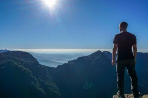 Ein Mann schaut von einem hohen Berg aus über eine nebelige Bergkette, die Sonne steht hoch am blauen Himmel.