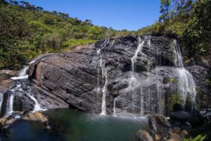 Kleiner Wasserfall mit Pool läuft über einen Felsen mit umgebender und mit Büschen bewachsener Hügellandschaft.