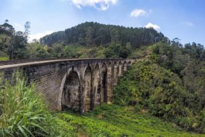 Alte Eisenbahnbrücke mit neun Bögen aus Stein zieht sich durch eine grüne und bewaldete Berglandschaft.