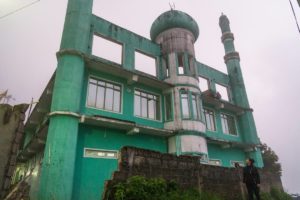 Farina steht vor einer türkisen, alten und verlassen wirkenden Moschee. Es ist nebelig.