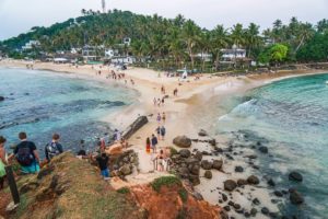 Touristen am Abend auf einem von Wasser umschlossenen Felsen im Meer, der durch eine Sandbank mit dem Strand verbunden ist. Im Hintergrund sind dichte Palmen und Hotels.
