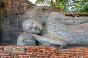 Aus dem Stein gehauene Statue eines liegenden und lächelnden Buddha hinter roter Steinmauer. Die hand ist unter den Kopf geschoben.