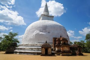 Eine kuppelförmige, weiße Stupa in historischen Ausgrabungsstätten vor blauem Himmel mit weißen Wolken, die Sonne brennt vom Himmel auf die trockene Landschaft.