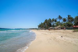 Palmengesäumter Strand mit Fischerbooten und türkis-blauem Meer vor wolkenlosem Himmel..