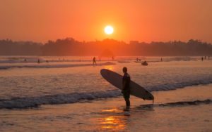 Ein Surfer steht mit Surfboard vor der untergehenden Sonne zum Sonnenuntergang im Meer während im Hintergrund weitere Surfer surfen