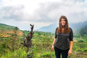 Farina steht vor einem großen Berghang in Ohiya über den sich Teefelder ziehen, im Hintergrund der Berge. Es ist bewölkt.