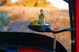 Eine kleine Buddha-Statue ist auf einen umgeklappten Spiegel in einem Tuk-Tuk montiert. Im Hintergrund sieht man eine große Wiese.