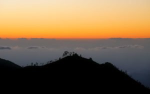 Morgengrauen - die Sonne geht auf vor den noch schwarzen Bergen im Vordergrund. Der mit Wolken besetzte Himmel färbt sich orangegelb und das Land dazwischen verschwimmt im blauen Nebel.