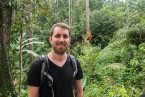 Steffen steht im Regenwald des Semenggho Wildlife Centre. Im Hintergrund hangeln zwei Orang-Utans an Seilen durch die Bäume.