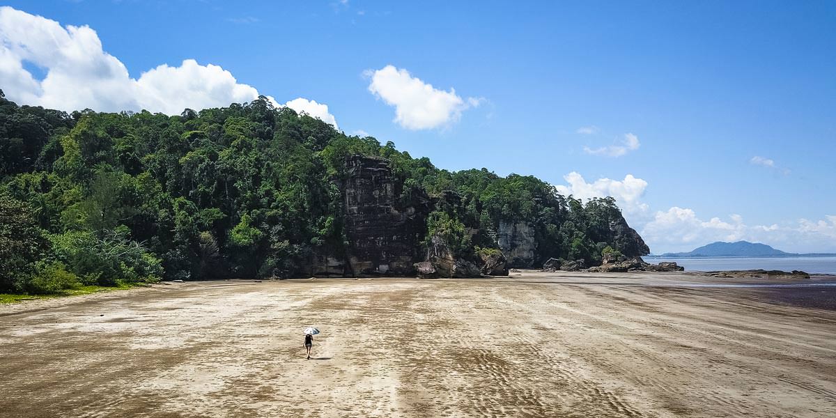 Farina geht an einem breiten, weitem und menschenleeren Strand, umgeben von Felsen und Regenwald mit einem Sonnenschirm spazieren.