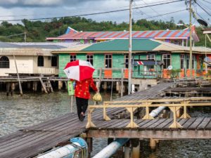 Eine Frau mit einem rot-weißen Schirm trägt Tüten über Holzwege zwischen Holzhäusern eines Wasserdorfes.