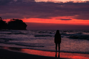 Farina steht in Miri am Strand. Der Sonnenuntergang leuchtet in rot und violett.