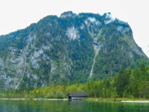 Eine kleine Hütte liegt am Königssee, im Hintergrund die Berge.